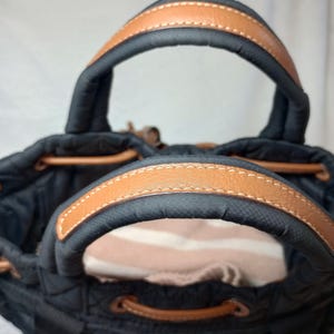 May include: Black tote bag with brown leather handles and trim. The bag has a drawstring closure and a quilted design. The handles are arched and stitched with white thread. The interior of the bag is visible.