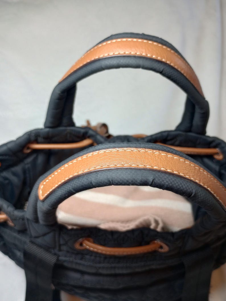 May include: Black tote bag with brown leather handles and trim. The bag has a drawstring closure and a quilted design. The handles are arched and stitched with white thread. The interior of the bag is visible.