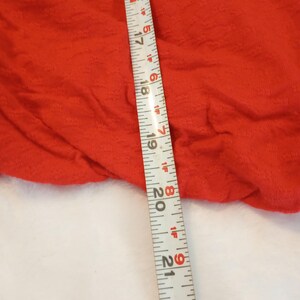 Puede incluir: Primer plano de una tela roja brillante y texturizada. Una cinta m&eacute;trica se coloca diagonalmente sobre la tela, mostrando medidas en pulgadas. La tela parece ser una prenda de vestir o textil.
