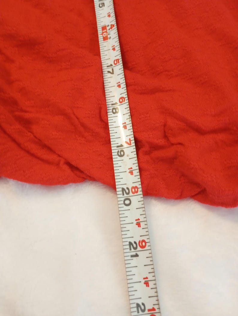 Puede incluir: Primer plano de una tela roja brillante y texturizada. Una cinta m&eacute;trica se coloca diagonalmente sobre la tela, mostrando medidas en pulgadas. La tela parece ser una prenda de vestir o textil.