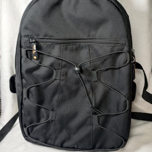 May include: A black backpack featuring a top handle, a horizontal zippered front pocket, and a black elastic bungee cord system with a central plastic toggle. The zipper pull on the front pocket displays the Amazon Basics logo in orange and white.