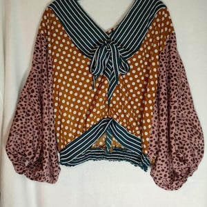 May include: A long-sleeve blouse featuring a mustard yellow body with white polka dots, teal and white striped trim at the neckline and hem, and pink sleeves with a dark brown animal print pattern. The blouse includes a tie detail at the front.