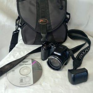 May include: A black Nikon Coolpix L310 digital camera with a lens cap, a black camera strap, a black carrying bag, and a Nikon User's Manual CD. The camera features a 21x wide optical zoom lens and is accompanied by its original accessories.