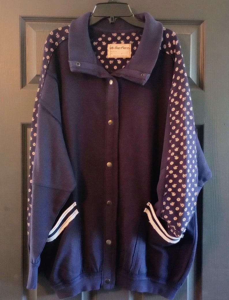 May include: A navy blue jacket with a button-up front, featuring white polka dot patterned sleeves and a ribbed collar. The jacket has a label that reads "We The Free". The jacket has two white stripes on the cuffs.