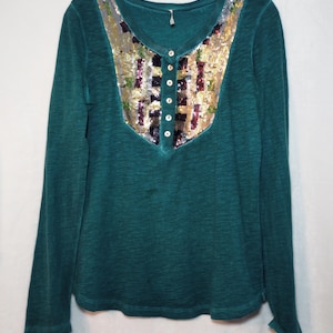 May include: Teal long-sleeve shirt with a sequined yoke. The yoke features a mix of gold, silver, and colorful sequins. The shirt has a Henley neckline with five small buttons. The fabric appears to be a soft, textured material.
