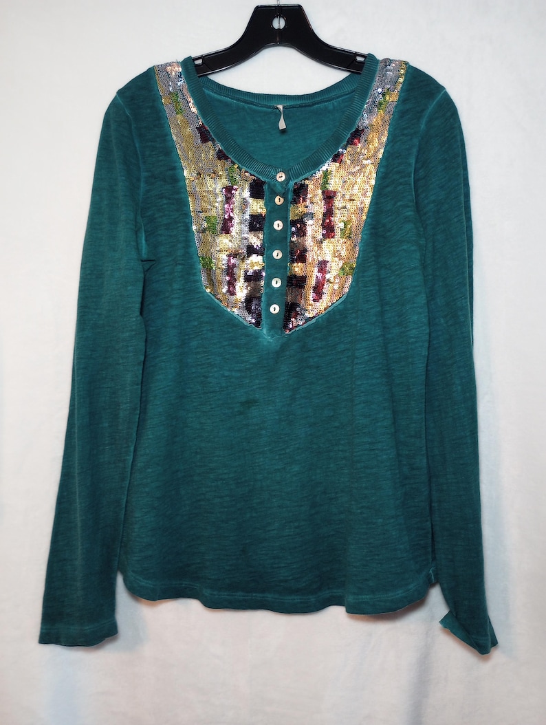 May include: Teal long-sleeve shirt with a sequined yoke. The yoke features a mix of gold, silver, and colorful sequins. The shirt has a Henley neckline with five small buttons. The fabric appears to be a soft, textured material.