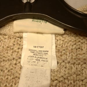 May include: A beige knitted sweater with a white care tag that reads "Machine wash warm water, no chlorine, tumble dry low, iron 110&deg;C, Laver &agrave; la machine &agrave; l'eau ti&egrave;de, pas de chlore, s&eacute;chage culbutage &agrave; basse temp&eacute;rature".