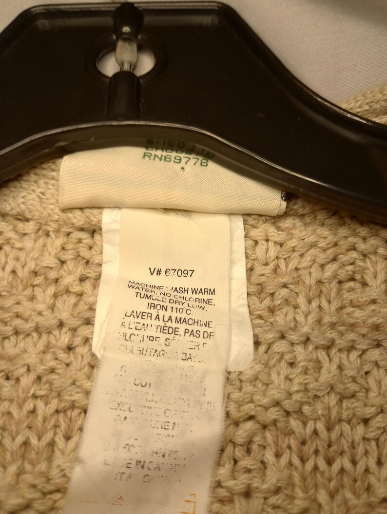 May include: A beige knitted sweater with a white care tag that reads "Machine wash warm water, no chlorine, tumble dry low, iron 110&deg;C, Laver &agrave; la machine &agrave; l'eau ti&egrave;de, pas de chlore, s&eacute;chage culbutage &agrave; basse temp&eacute;rature".
