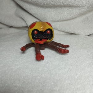 May include: A yellow and red toy creature with a round head and multiple brown legs. The creature has red eyes and a red tongue. The toy is on a white surface.