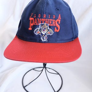 Puede incluir: Gorra de béisbol azul marino y roja con las palabras "FLORIDA PANTHERS" en naranja. La gorra tiene una visera roja y un logotipo de pantera en el centro.