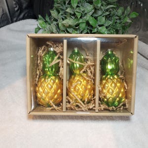 May include: Three glass pineapple ornaments in a cardboard box. The ornaments have a gold body and green top, with a textured surface. The box is filled with shredded paper for protection. A decorative item for the holidays.