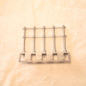 May include: A silver-colored, rectangular architectural element with vertical bars and a horizontal top. The base has five rectangular openings with a V-shaped detail. The design suggests a miniature fence or gate.