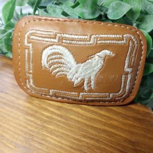 May include: A rectangular tan leather belt buckle featuring a cream-colored embroidered rooster in the center, surrounded by a geometric Greek key border pattern stitched in the same cream thread along the perimeter of the leather surface.