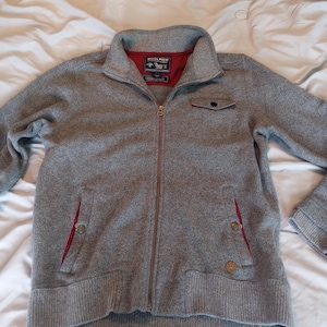May include: A gray zip-up jacket with a high collar. The jacket has a red lining visible at the collar and pocket edges. It features two front pockets and a small chest pocket. The jacket has a ribbed hem and cuffs.
