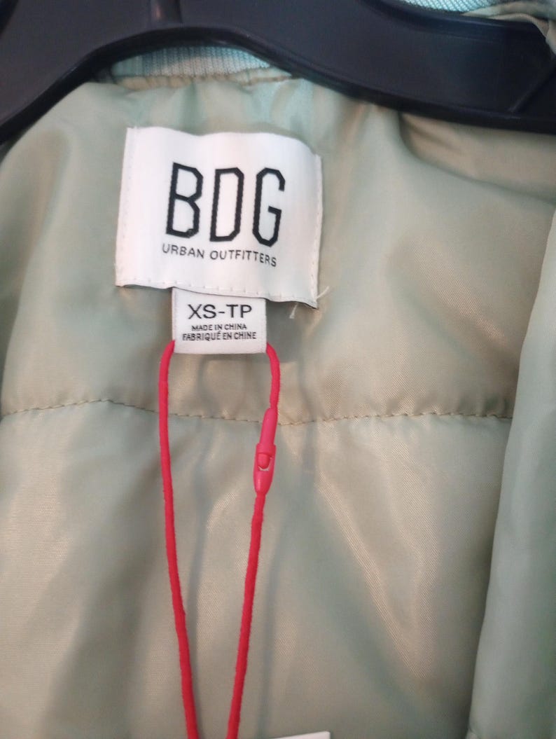 Może przedstawiać: Zbliżenie metki odzieżowej marki BDG firmy Urban Outfitters. Biała metka zawiera czarny tekst, w tym nazwę marki i rozmiar XS-TP. Dołączony jest jasnoczerwony sznurek.