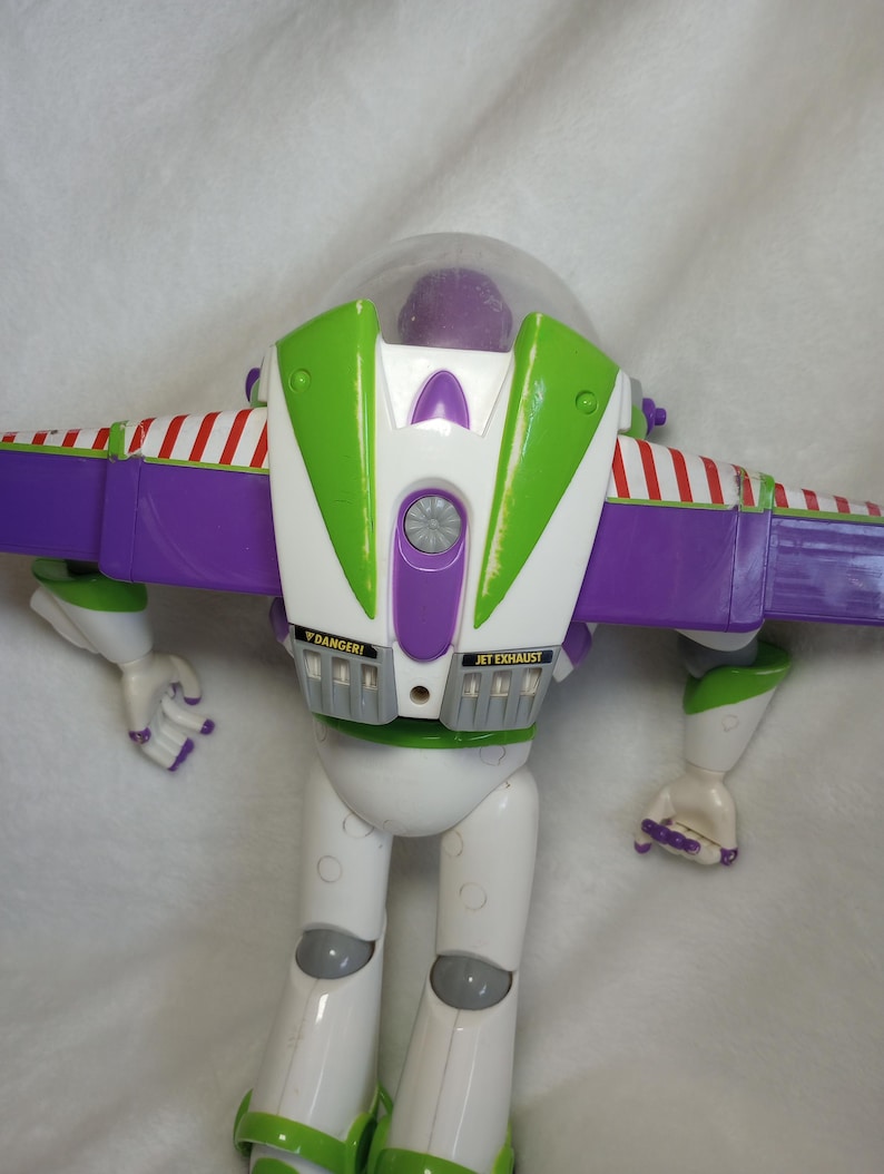 May include: A plastic action figure of a space ranger character featuring a white, purple, and lime green color scheme. The figure has a clear dome helmet, extendable purple wings with red and white striped accents, and labels reading DANGER and JET EXHAUST.