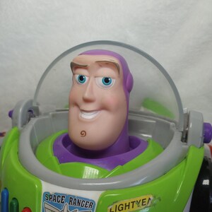 May include: A Buzz Lightyear action figure featuring a purple hood, a clear plastic helmet visor, and a white chest plate with green accents. The chest displays a Space Ranger emblem and the text LIGHTYEAR in black letters on a yellow background.