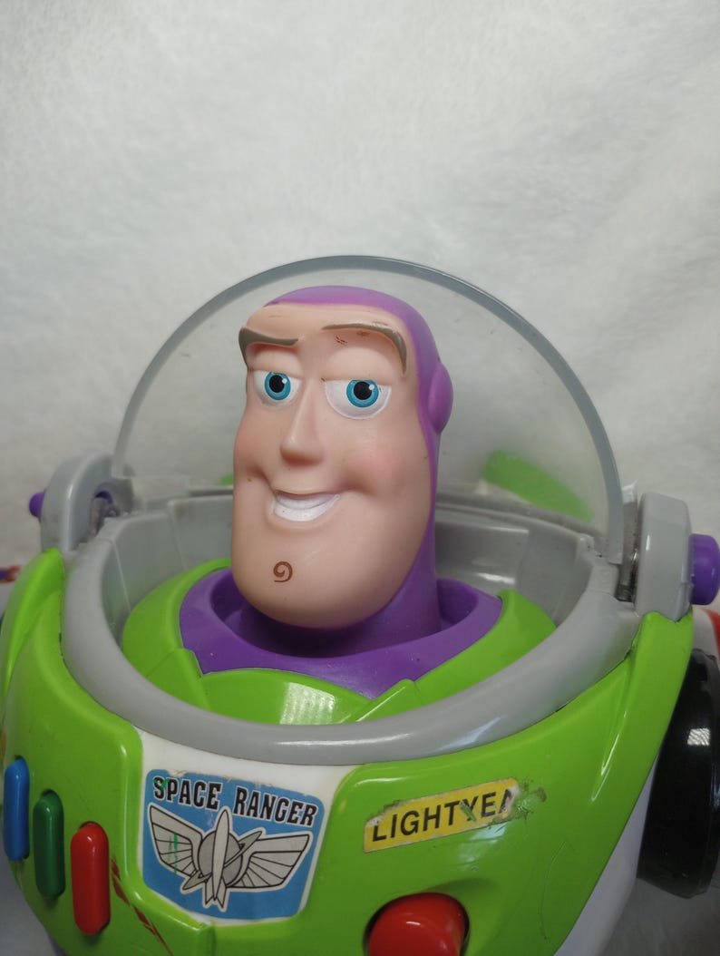 May include: A Buzz Lightyear action figure featuring a purple hood, a clear plastic helmet visor, and a white chest plate with green accents. The chest displays a Space Ranger emblem and the text LIGHTYEAR in black letters on a yellow background.
