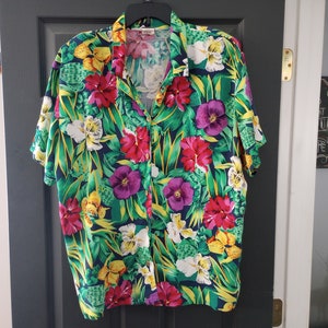 Puede incluir: Una camisa de manga corta con botones y estampado tropical con flores grandes en tonos rojos, amarillos, morados y blancos sobre un fondo azul oscuro.