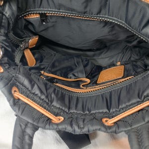 May include: Black quilted nylon backpack with brown leather accents. The interior features multiple compartments and a zippered pocket. The bag has brown drawstrings and a black shoulder strap. The bag is open, revealing the interior.