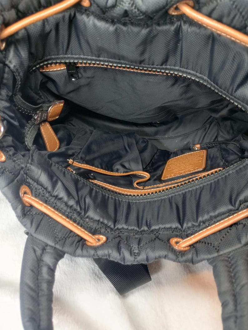 May include: Black quilted nylon backpack with brown leather accents. The interior features multiple compartments and a zippered pocket. The bag has brown drawstrings and a black shoulder strap. The bag is open, revealing the interior.