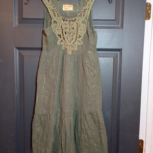 K&ouml;nnte beinhalten: Olivgr&uuml;nes &auml;rmelloses Kleid mit dekorativem Spitzenausschnitt und goldenen Metallic-Faden-Akzenten. Das Kleid hat einen Stufenrock und h&auml;ngt an einem schwarzen Kleiderb&uuml;gel. Das Kleid ist aus leichtem Stoff gefertigt.