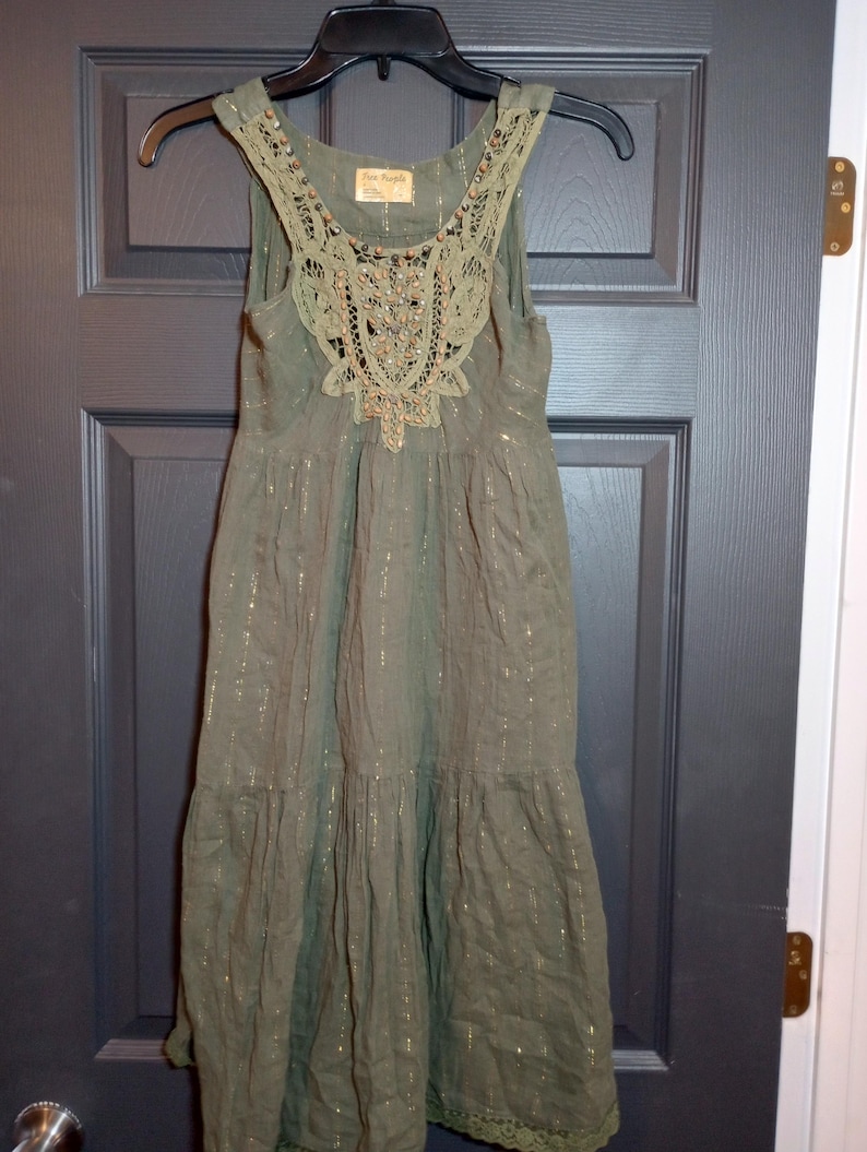 K&ouml;nnte beinhalten: Olivgr&uuml;nes &auml;rmelloses Kleid mit dekorativem Spitzenausschnitt und goldenen Metallic-Faden-Akzenten. Das Kleid hat einen Stufenrock und h&auml;ngt an einem schwarzen Kleiderb&uuml;gel. Das Kleid ist aus leichtem Stoff gefertigt.