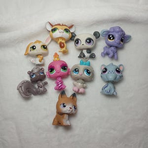 May include: A collection of nine small plastic animal figurines featuring stylized designs with large eyes. The set includes a brown and white deer, a panda, a purple lamb, a tan rabbit, a pink creature, a gray bird, a squirrel, a cat, and a fox.