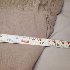 May include: A light brown garment with a lace detail at the top. The garment is measured with a tape measure, showing a width of approximately 17 inches.