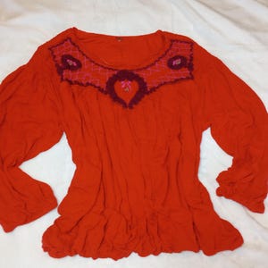 Puede incluir: Una blusa roja de manga larga con dobladillo con volantes. El cuello redondo est&aacute; adornado con un dise&ntilde;o bordado en burdeos y rosa. Las mangas est&aacute;n recogidas en los pu&ntilde;os. La blusa est&aacute; hecha de una tela suave y fluida.