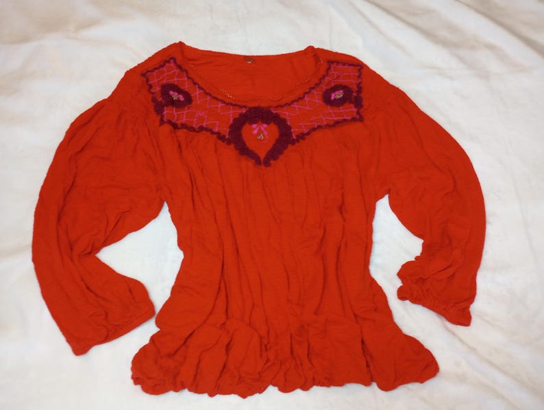 Puede incluir: Una blusa roja de manga larga con dobladillo con volantes. El cuello redondo est&aacute; adornado con un dise&ntilde;o bordado en burdeos y rosa. Las mangas est&aacute;n recogidas en los pu&ntilde;os. La blusa est&aacute; hecha de una tela suave y fluida.