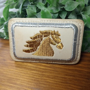 May include: A rectangular off-white belt buckle featuring a central embroidered gold horse head profile. The design is framed by a metallic silver stitched border, with visible stitching along the outer edge of the cream-colored material.