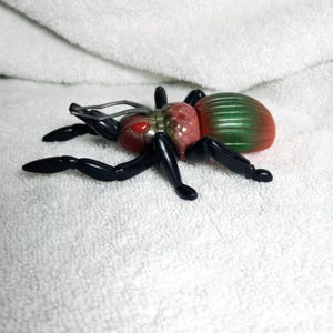 May include: A plastic toy insect with a red head, a segmented body that transitions from green to red, and black legs. The insect has large red eyes and silver antennae. The toy is set against a white background.