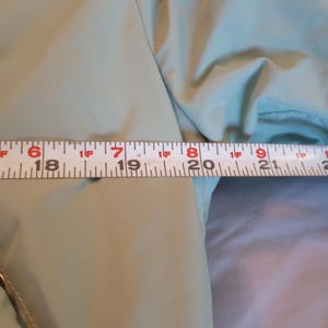 Może przedstawiać: Zbliżenie na jasny zielony rękaw kurtki z taśmą mierniczą. Taśma wskazuje pomiar około 48 cm. Kurtka ma gładką, lekko błyszczącą fakturę.