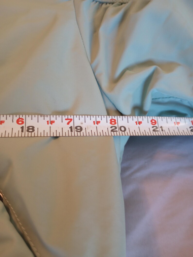 Może przedstawiać: Zbliżenie na jasny zielony rękaw kurtki z taśmą mierniczą. Taśma wskazuje pomiar około 48 cm. Kurtka ma gładką, lekko błyszczącą fakturę.
