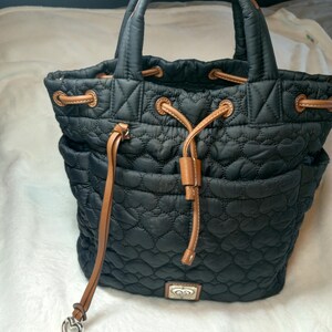 May include: Black quilted tote bag with a heart pattern. Features brown leather accents on the drawstring closure and handles. Includes a silver heart-shaped charm and a decorative metal emblem.