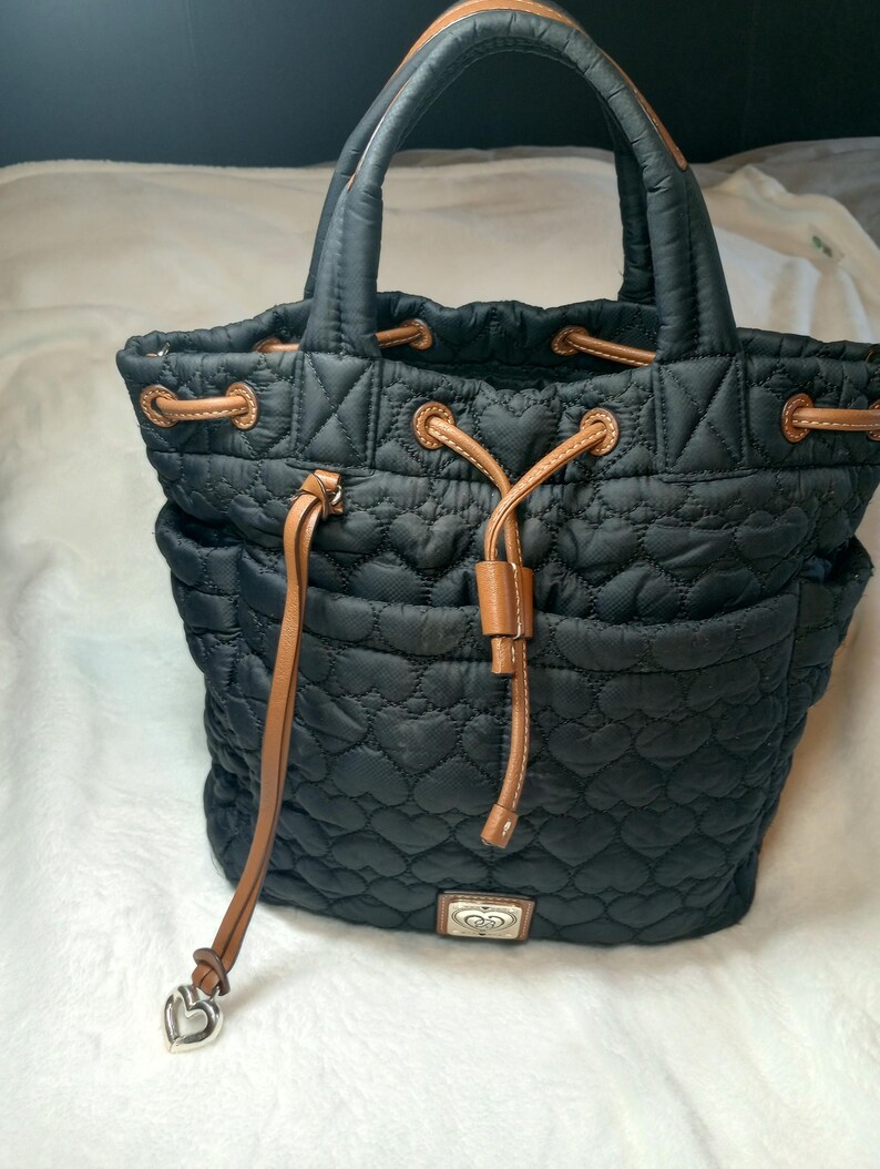 May include: Black quilted tote bag with a heart pattern. Features brown leather accents on the drawstring closure and handles. Includes a silver heart-shaped charm and a decorative metal emblem.