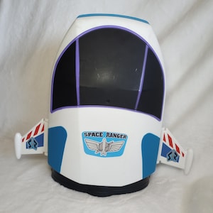 Op de afbeelding: Een wit plastic Space Ranger speelgoedruimteschip met een grote zwarte voorruit, blauwe accenten en twee zijvleugels met rood-witte strepen. De voorkant toont een blauw embleem met de tekst SPACE RANGER en een afbeelding van een gevleugelde raket.