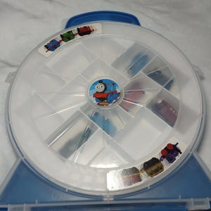 以下が含まれることがあります： 青と白のプラスチック製トーマス・ザ・タンク・エンジンの玩具収納ケース。ケースは丸く、内部は仕切られており、青いハンドルが付いています。蓋にはトーマス・ザ・タンク・エンジンのグラフィックと、さまざまな車両のステッカーが付いています。