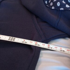 May include: Navy blue sweatshirt with a white floral pattern on the shoulder. The tape measure shows a length of approximately 26 inches.