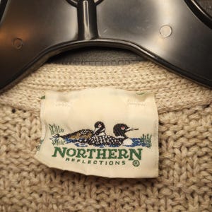 May include: A beige knitted sweater with a label that reads "Northern Reflections" and features two loon birds in a lake setting.