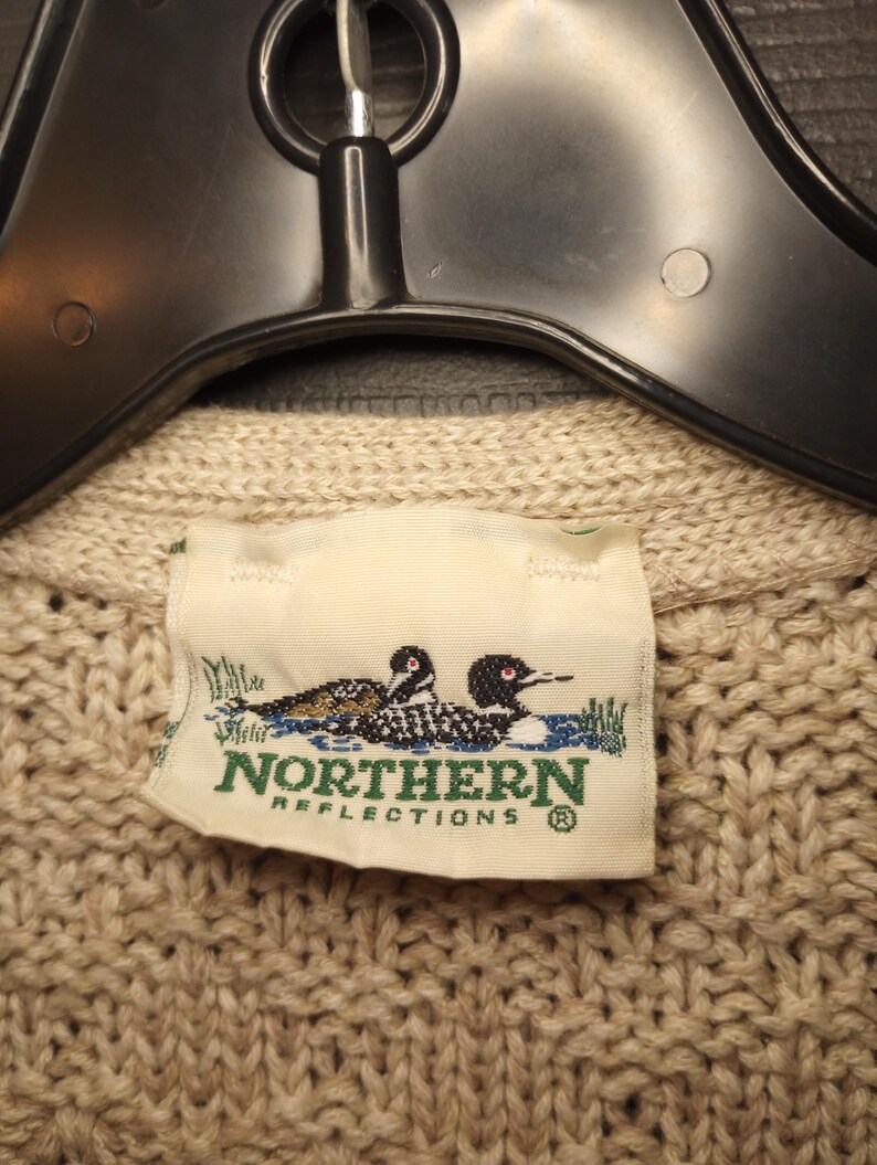 May include: A beige knitted sweater with a label that reads "Northern Reflections" and features two loon birds in a lake setting.