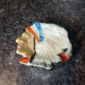 May include: A ceramic ashtray shaped like a Native American chief's head. The ashtray features a detailed face with a headdress in white, red, blue, and black. The piece is likely vintage and has a glossy finish.