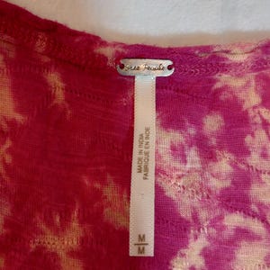 May include: A close-up of a pink and beige tie-dye top. The top has a scoop neck and a tag that reads "Free People". The tag also says "Made in India Fabriqu&eacute; en Inde" and has a size "M".