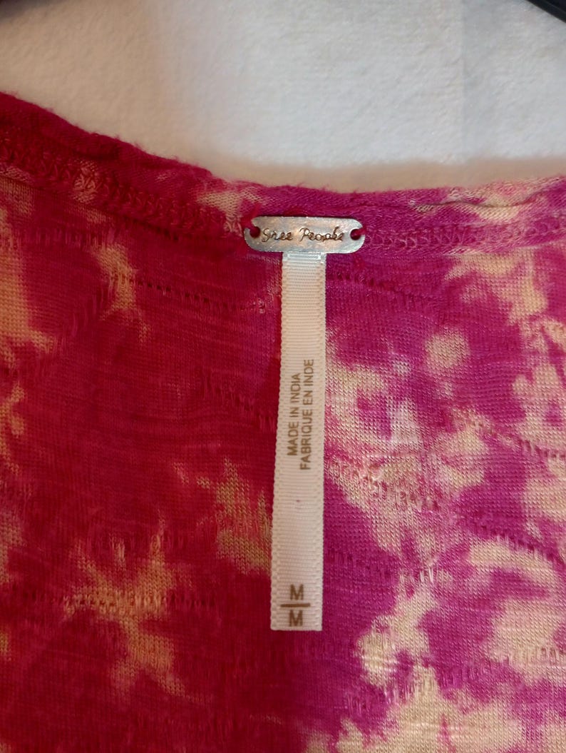 May include: A close-up of a pink and beige tie-dye top. The top has a scoop neck and a tag that reads "Free People". The tag also says "Made in India Fabriqu&eacute; en Inde" and has a size "M".