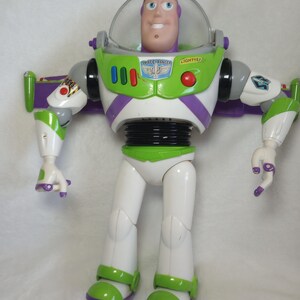 May include: A Buzz Lightyear action figure featuring a white, lime green, and purple plastic suit with a clear retractable helmet. The chest displays the text SPACE RANGER and LIGHTYEAR with a blue and silver emblem, three red buttons, and a red circular button.