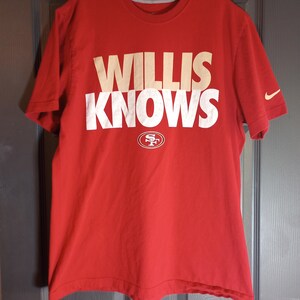 May include: A red t-shirt with the words "WILLIS KNOWS" in large white and tan letters. Below the text is the San Francisco 49ers logo. A Nike logo is visible on the sleeve.