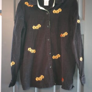 May include: A black long-sleeved button-down shirt with embroidered pumpkin faces. The shirt has a light green lining around the collar.