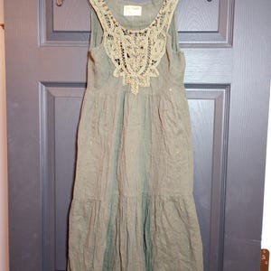 K&ouml;nnte beinhalten: Olivgr&uuml;nes &auml;rmelloses Kleid mit einem mit Spitze und Pailletten verzierten Oberteil. Das Kleid hat einen Stufenrock und h&auml;ngt an einem schwarzen Kleiderb&uuml;gel. Das Kleid hat einen dezenten Schimmer und eine Spitzenborte am Saum.