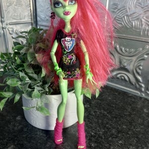 Peut inclure: Une poupée Monster High avec une peau verte, des cheveux roses et des yeux bleus. La poupée porte un haut noir avec un emblème de crâne et une jupe à motifs, des bottes roses et des accessoires verts. Un pot blanc avec une plante verte est en arrière-plan.